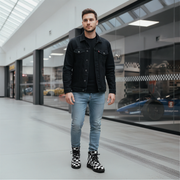 Man wearing a black denim jacket, light blue jeans, and checkered boots standing in a modern indoor setting.