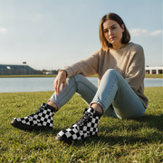 Woman sitting on grass wearing black and white checkered boots, beige sweater, and light blue jeans.