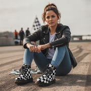 Woman sitting on a racetrack wearing checkered boots, leather jacket, and jeans.