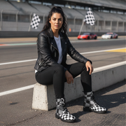Woman in black leather jacket and checkered boots sitting on a concrete block at a race track.