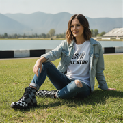 Woman sitting on grass wearing a denim jacket and white t-shirt with text, and checkered boots,  mountains in the background