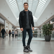 Man standing in a modern mall wearing a black jacket, gray shirt, blue jeans, and checkered boots.