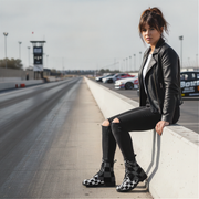 Woman in black leather jacket and checkered boots sitting on a race track.