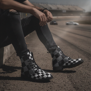 Man wearing checkered boots sitting on a race track with cars in the background
