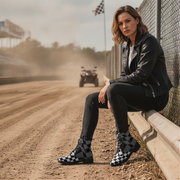 Woman wearing checkered boots sitting on a guardrail by a dirt track with a blurred ATV in the background