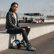 Woman sitting on a race track with a helmet and checkered boots, with a race car in the background.