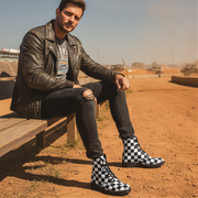 Man sitting on a wooden bench wearing checkered boots in an outdoor setting.