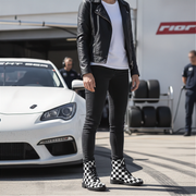 Person wearing a black leather jacket, white shirt, black pants, and checkered boots standing in front of a white car.