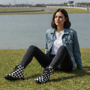 Woman sitting on grass by a water body wearing checkered boots, denim jacket, and black pants.