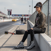 Woman sitting on a race track barrier wearing checkered boots, with a race car and track in the background.