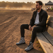 Man sitting on a wooden bench at a dirt track with motorcycles in the background and wearing checkered boots