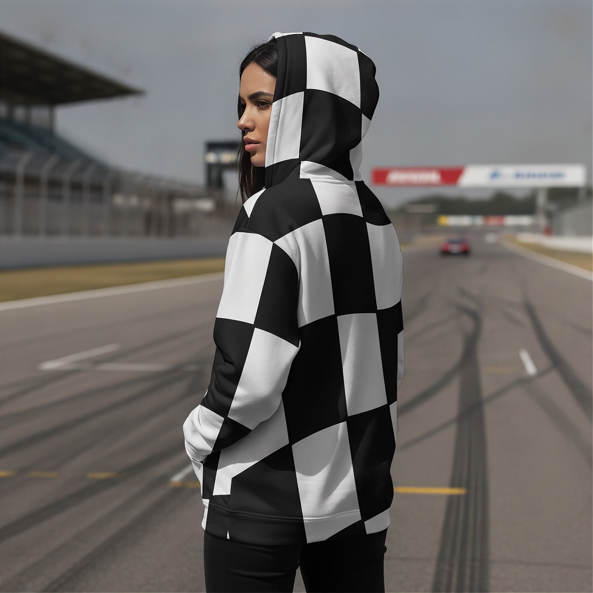A woman wearing a black and white checkered hoodie stands on a racetrack, looking over her shoulder. The background shows a blurred racetrack with a race car and grandstands.
