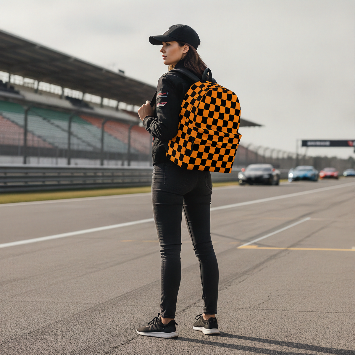 A woman standing on a racetrack wearing a black cap, black jacket, black skinny jeans, and black sneakers, carrying a black and orange checkered backpack.