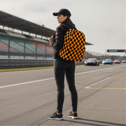 A woman standing on a racetrack wearing a black cap, black jacket, black skinny jeans, and black sneakers, carrying a black and orange checkered backpack.