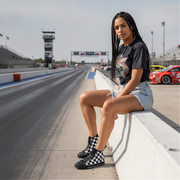 Woman wearing checkered shoes sitting on a race track barrier with cars in the background