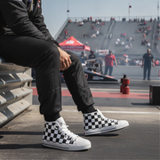 Man wearing checkered sneakers sitting on a race track with a race car and grandstand in the background.