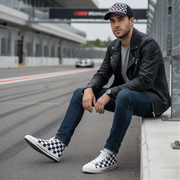 Man sitting on a race track wearing a black leather jacket, jeans, and checkered shoes.