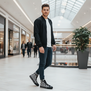 Man standing in a modern mall wearing a black jacket, white shirt, blue jeans, and checkered shoes.