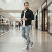Man in a black jacket, white shirt, light blue jeans, and checkered sneakers standing in a mall.
