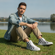 Man sitting on grass wearing a denim jacket, beige pants, and checkered shoes with a lake and mountains in the background.