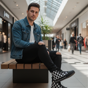 Man sitting on a bench in a shopping mall wearing a denim jacket and checkered shoes.