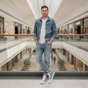 Man in a denim jacket and jeans and wearing checkered shoes standing in a modern mall.