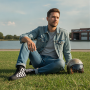 Man sitting on grass with a helmet next to him, wearing a denim jacket and jeans and checkered high top shoes.