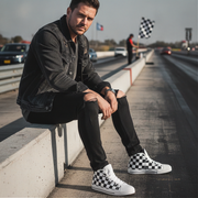 Man sitting on a concrete barrier at a race track wearing checkered shoes.