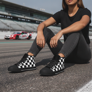 Woman wearing black and white checkered shoes sitting on a race track with racing cars in the background.