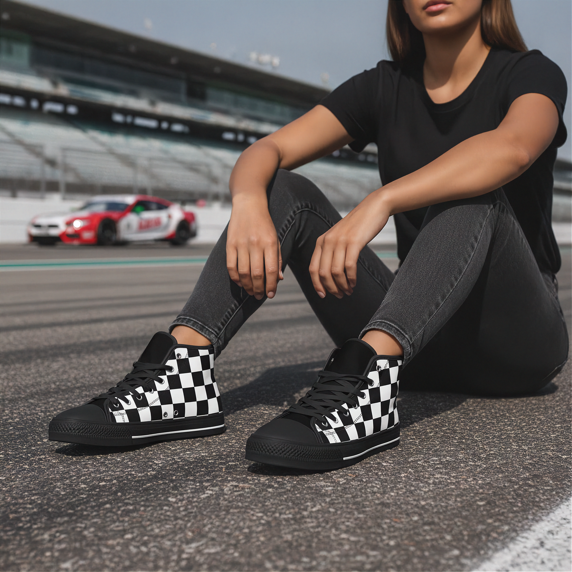 Woman wearing black and white checkered shoes sitting on a race track with racing cars in the background.