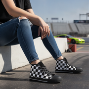 Man wearing checkered black and white high top shoes sitting on a concrete barrier with a race track in the background.