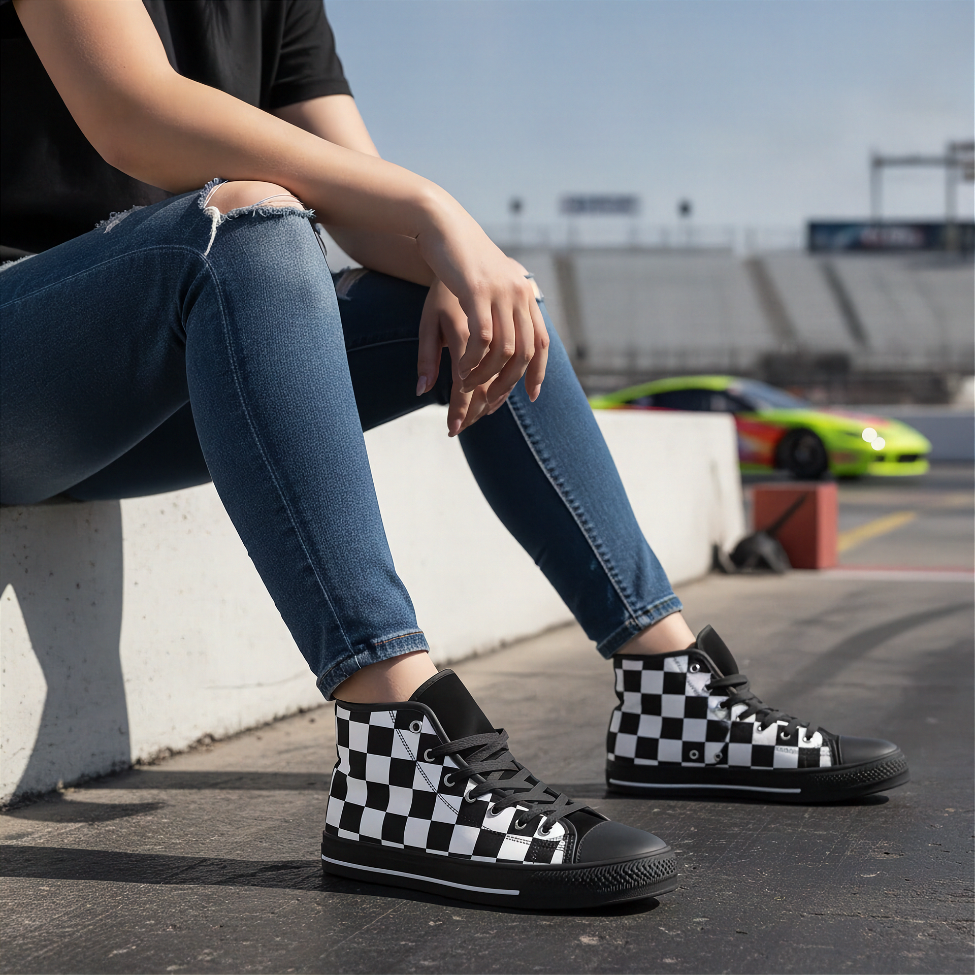 Man wearing checkered black and white high top shoes sitting on a concrete barrier with a race track in the background.