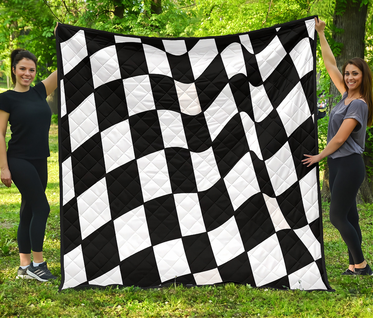 A large black and white checkered quilt is held up outdoors by two women standing on grass in front of trees.