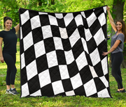 A large black and white checkered quilt is held up outdoors by two women standing on grass in front of trees.