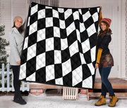 Two women holding up a large quilted blanket featuring a black and white checkered flag pattern, set against a snowy porch background.