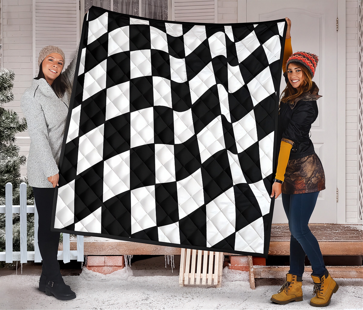 Two women holding up a large quilted blanket featuring a black and white checkered flag pattern, set against a snowy porch background.