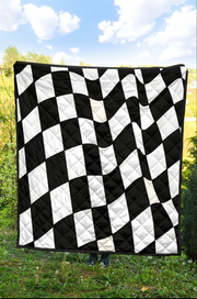 A black and white checkered quilted blanket or throw is held up outdoors in a grassy field under a blue sky with clouds.