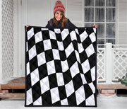 A woman wearing a red knit beanie holds up a large quilted blanket with a black and white wavy checkerboard pattern in front of a snowy porch.