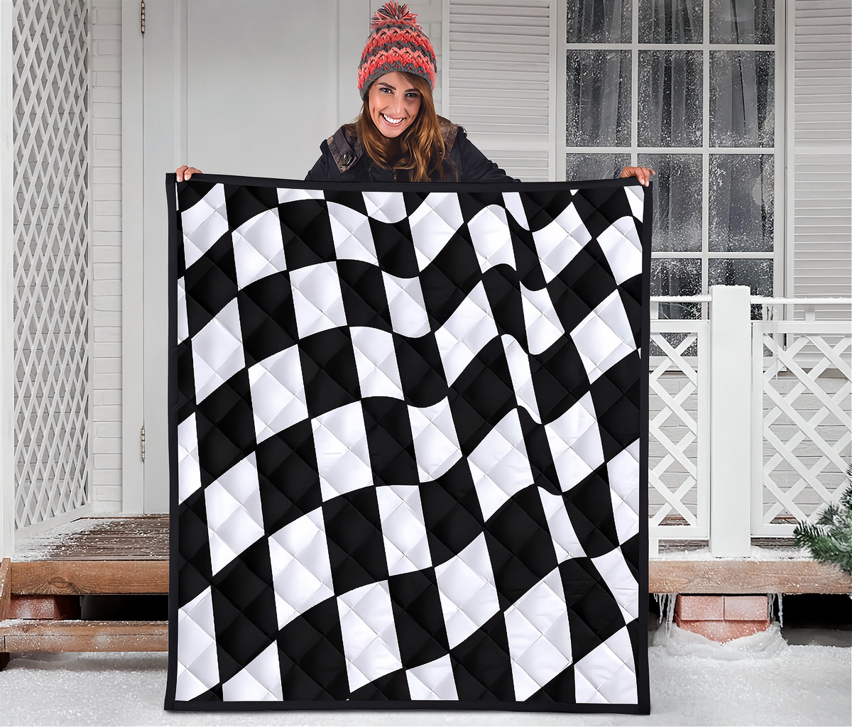 A woman wearing a red knit beanie holds up a large quilted blanket with a black and white wavy checkerboard pattern in front of a snowy porch.