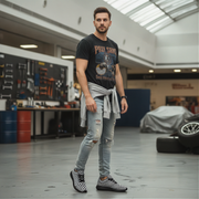 Man standing in a garage wearing a black t-shirt with graphic design and ripped jeans and checkered shoes