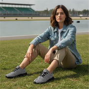 Woman sitting on grass wearing checkered shoes with a lake and stadium in the background