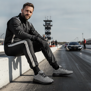 Man sitting on a race track with checkered shoes, cars in the background
