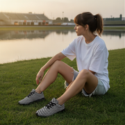 Woman sitting on grass wearing checkered shoes with a lake and stadium in the background