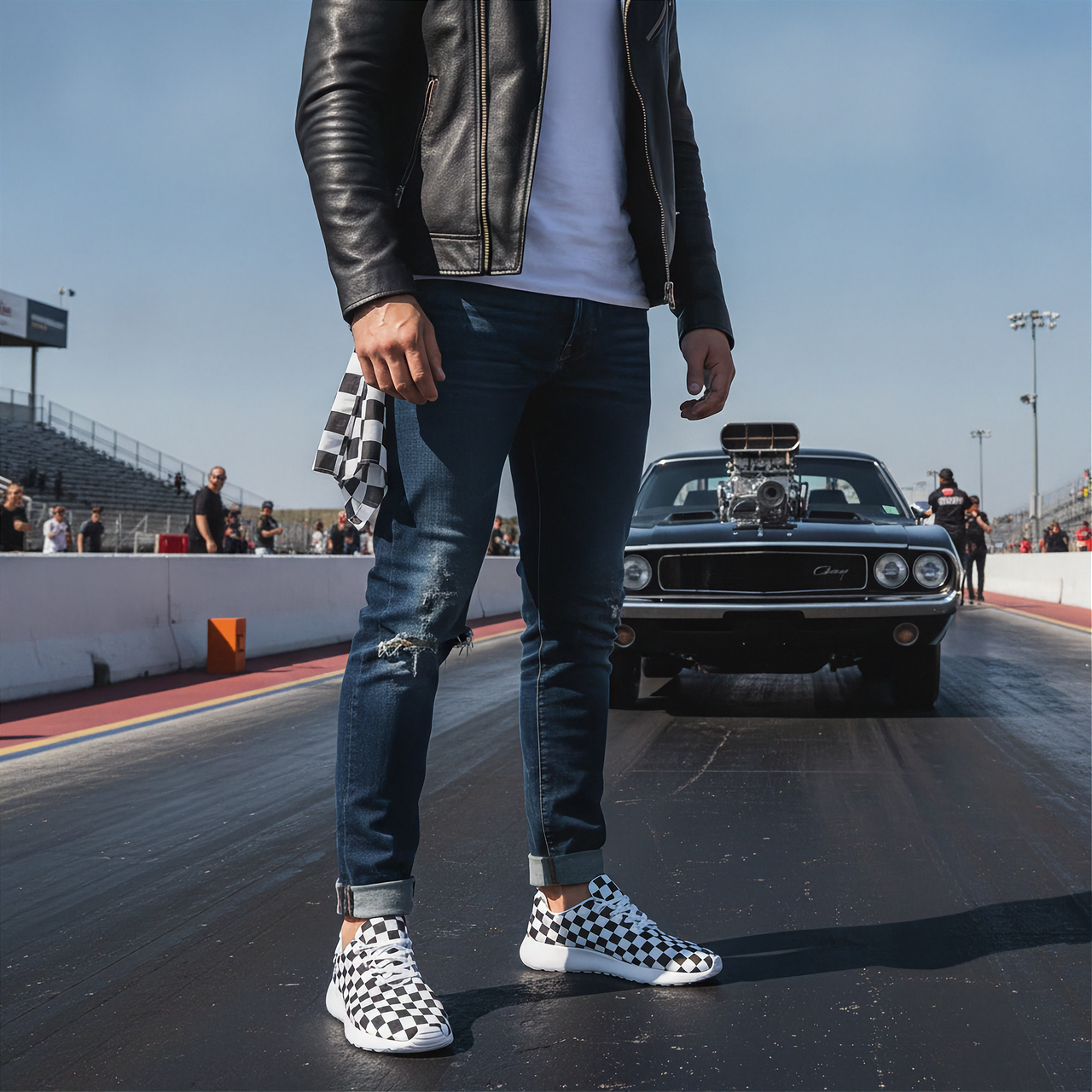 Man wearing a leather jacket, jeans, and checkered shoes on a racetrack with a car in the background.
