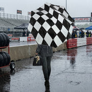 A person walking in the rain at a racetrack holding a large black-and-white checkered umbrella. The racetrack, wet pavement, tires, and trackside barriers are visible in the background.