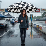 A woman stands on a wet racetrack holding a large black-and-white checkered umbrella. She wears a dark rain jacket, dark leggings, and black boots.