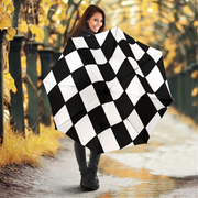 A woman stands on a wet street under trees holding a large black-and-white checkered umbrella. She wears a dark rain jacket, dark leggings, and black boots.