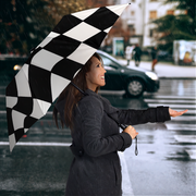 A woman in a dark gray coat stands in the rain on a city street holding a large black-and-white checkered umbrella, with a blurred car and city background behind her.