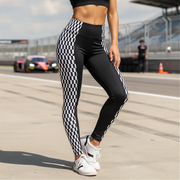 A person standing on a racetrack wearing black leggings with a black-and-white checkered side panel and white sneakers. A blurred race car and track barriers are visible in the background.