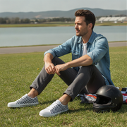 Man sitting on grass with a helmet, wearing a denim jacket and checkered shoes, with a scenic background.